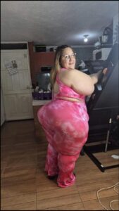 A woman wearing a pink tie-dye jumpsuit stands in a kitchen, leaning against a piece of exercise equipment. She has large hips and thighs, and is wearing glasses.