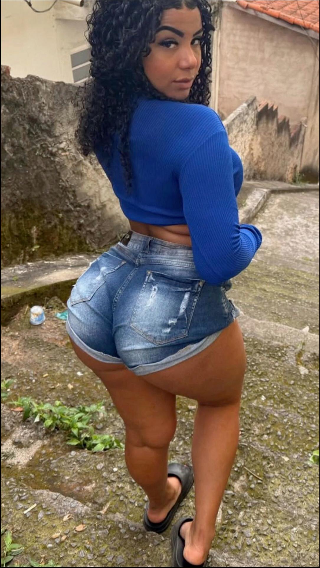 A woman with curly dark hair wears a blue cropped top and distressed denim shorts. She stands on a stone surface with vegetation and a wall in the background.