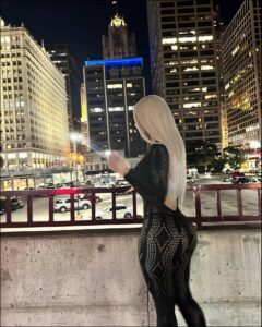 A woman with long blonde hair, wearing a black patterned bodysuit, stands on a concrete ledge at night, facing a cityscape with illuminated buildings and a prominent tower. Cars are visible on the street below.