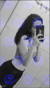 A young woman takes a mirror selfie in a bathroom, wearing a patterned top and dark bottoms. A repeating smiley face pattern is overlaid on the black and white image.