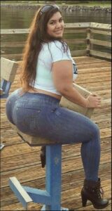 A woman with long brown hair sits on a blue metal stool on a wooden pier. She wears a white t-shirt, blue jeans, and black ankle boots. A wooden railing and body of water are visible in the background.