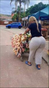 A woman pushes a shopping cart filled with produce, including potatoes, onions, and leafy greens, down a paved street. A blue car is parked in the background, and a blue canopy is visible to the right.