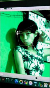 A selfie taken on a green-lit screen displays a person wearing a tropical-print shirt. Two skeletal figures are superimposed on the image, one near the person's head and the other lower down. The screen's interface shows various application icons at the bottom.