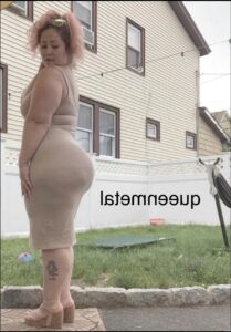 A woman with pink hair, wearing a beige dress and sandals, stands with her back to the camera in front of a house. She has a tattoo on her left leg and wears sunglasses perched on her head. The house has light-colored siding and multiple windows. A green lawn, stone border, and a hammock are visible in the background.