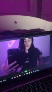 A person takes a selfie on a laptop screen, holding the phone in front of their face. The laptop screen displays a purple background and a partially visible image. The laptop's keyboard and trackpad are visible below the screen.