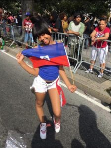 A young woman holds a Haitian flag on a city street, smiling at the camera. She wears a blue tank top, white shorts, and pink and white sneakers. Metal barriers line the street behind her, with onlookers standing behind them. Trees and buildings are visible in the background.