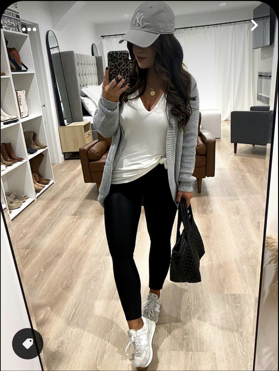 A woman takes a mirror selfie in a room. She wears a gray NYC baseball cap, a white v-neck top, black leggings, and white sneakers. She carries a black quilted handbag and holds a leopard-print phone in her right hand. A partial view of a closet with shoes, a bed, and a brown leather chair is visible in the background.