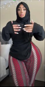 A woman wearing a black hijab, black long-sleeved top, and patterned leggings stands with her hands clasped in front of her chest. She is posing against a wall with string lights and a portion of a bed is visible in the background.