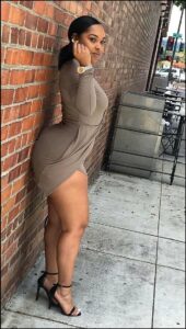 A woman in a light brown, form-fitting mini dress and black high-heeled sandals stands against a brick wall. She wears a gold watch and earrings, and her hair is pulled back. She is positioned near a metal planter and outdoor seating area.