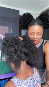 Two young girls with styled black curly hair. One girl, wearing a floral top, stands behind the other girl, who is adjusting her hair. They are in a room with dark walls and a partially visible green object.